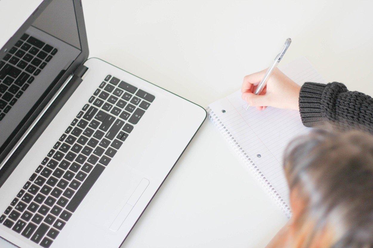 Person writing on a note pad next to an open laptop computer